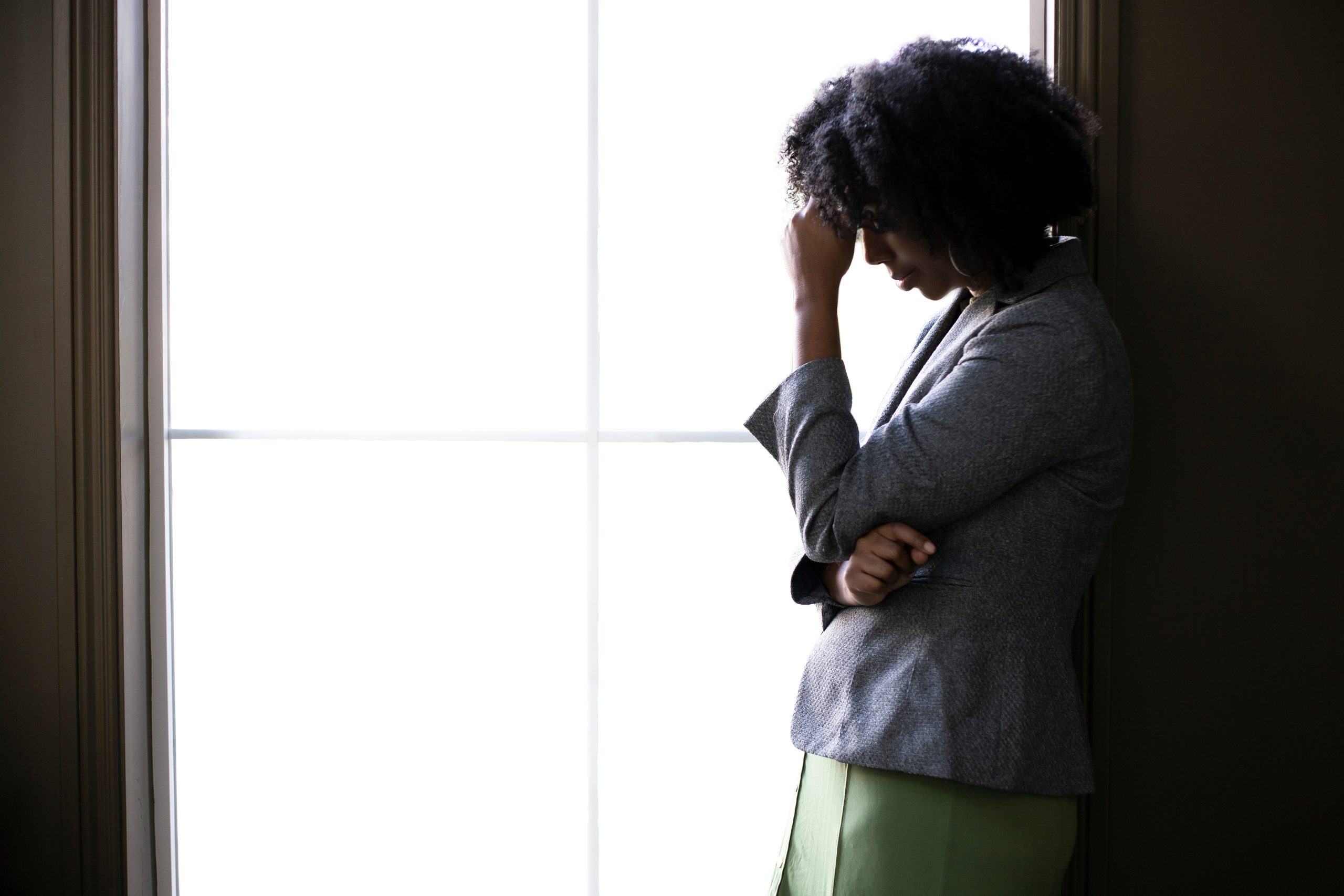 Silhouette of a stressed out black African American businesswoman looking worried and thinking about problems and failure by the office window.  She looks depressed or upset about debt or bankruptcy.